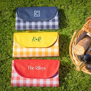 Puede incluir: Tres mantas de picnic personalizadas en azul marino, amarillo y rojo con estampados de cuadros. La manta azul marino tiene una inicial blanca, la manta amarilla tiene iniciales y la manta roja tiene un apellido. Una cesta de picnic de mimbre est&aacute; llena de comida y bebida.