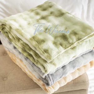 May include: A stack of four plush blankets in shades of green, gray, and cream. The top blanket has a tie-dye pattern. The blankets are folded neatly and stacked on top of each other. The text "The Warners" is embroidered on the top blanket.