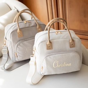 May include: Two light gray corduroy baby bags with tan handles and gold hardware. One bag has the name "Amelia" embroidered, the other "Christina". Both have a shoulder strap and a front pocket.
