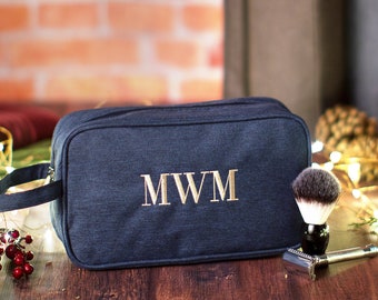 May include: A dark blue toiletry bag with a zipper closure and a handle. The bag has the initials "MWM" embroidered on the front in gold thread. The bag is sitting on a wooden surface with a shaving brush and razor nearby.