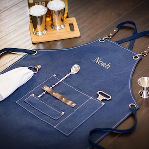 May include: A blue denim bar apron with a brown leather strap and a large pocket. The apron has a name embroidered on it, "Noah". The apron is laying on a wooden surface with a cocktail shaker, two jiggers, a bar spoon, and a bottle opener.