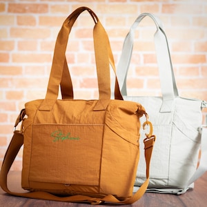 May include: Two tote bags. One is a mustard yellow color with a shoulder strap and the name "Stephanie" embroidered in green. The other is a light gray color with a shoulder strap. Both bags have top handles.