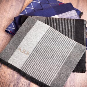 May include: A stack of folded scarves in various patterns and colors. The top scarf is gray with vertical stripes and the initials "A.R.S." embroidered on it. Other scarves include blue diamond and plaid patterns. The scarves are on a wooden surface.