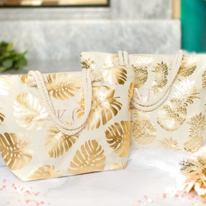 May include: Two white canvas tote bags with gold foil tropical leaf and pineapple designs. The bags have rope handles and are personalized with the initials "K" and "M".