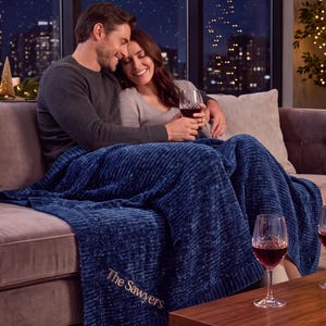 May include: A deep blue, textured, plush throw blanket featuring the embroidered text 'The Sawyers' in a gold-toned script font. The blanket has a ribbed border and a soft, velvety appearance, draped over a sofa in a cozy indoor setting.