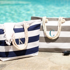 May include: Two striped canvas tote bags with rope handles. The bag on the left is navy blue and white with a gold 'K' monogram. The bag on the right is gray and white with a teal 'C' monogram.