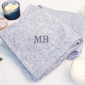 May include: A gray and white throw blanket with the initials "MB" embroidered in dark gray. The blanket is folded and displayed on a white surface with two candles.