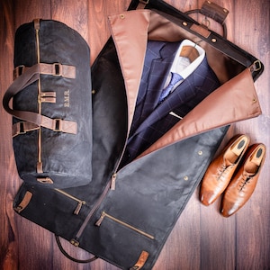 May include: A black canvas garment bag with a brown leather trim and a zipper closure. The bag is open and a suit jacket, a white dress shirt, and a tie are inside. Two brown leather shoes are also visible.