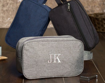May include: Three toiletry bags in different colors: navy blue, black, and gray. The gray bag has the initials "JK" embroidered on the front.