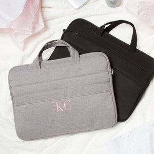 May include: Two laptop sleeves, one gray with the initials "KC" embroidered in pink and one black. Both sleeves have a handle and are made of a soft, textured fabric.