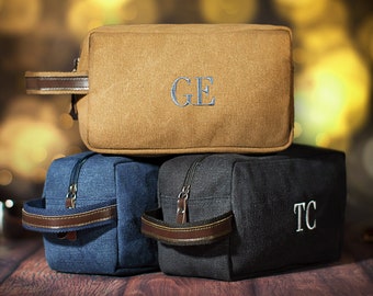 May include: Three personalized toiletry bags in tan, blue, and black. Each bag has a zipper closure, a brown leather handle, and embroidered initials. The tan bag has "GE", the black bag has "TC", and the blue bag is unadorned.