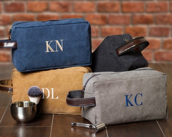 May include: Four canvas toiletry bags with leather handles. The bags are in different colors: blue, brown, gray, and black. Each bag has a different monogram embroidered on the front: KN, DL, KC, and a fourth bag with no monogram.