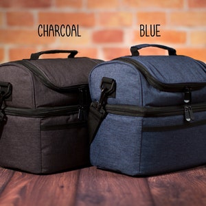 May include: Two insulated lunch bags, one charcoal gray and one blue, with double zippers and a shoulder strap. The bags are sitting on a wooden surface.