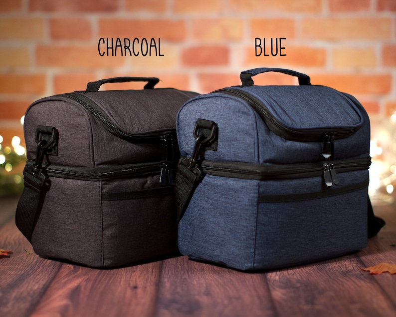 May include: Two insulated lunch bags, one charcoal gray and one blue, with double zippers and a shoulder strap. The bags are sitting on a wooden surface.