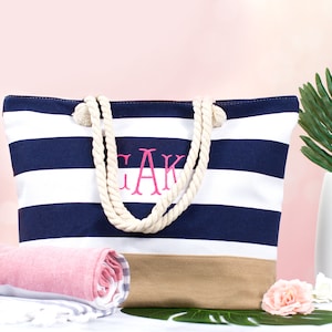 May include: A navy and white striped tote bag with rope handles and a tan bottom. The bag features embroidered pink initials. A rolled pink and white towel and a white vase with a green leaf are in the background.