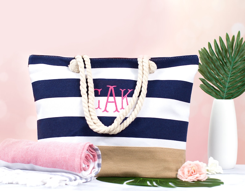 May include: A navy and white striped tote bag with rope handles and a tan bottom. The bag features embroidered pink initials. A rolled pink and white towel and a white vase with a green leaf are in the background.