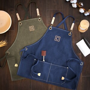 May include: Two canvas aprons with adjustable straps and pockets. One apron is olive green with the letter "R" embroidered on the front. The other apron is navy blue with the letter "C" embroidered on the front.