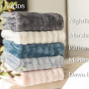 May include: A stack of six plush blankets in various solid colors. The colors include Nightfall, Marshmallow, Patina, Morning Mist, and Dawn Blush. The blankets are soft and fluffy, perfect for adding warmth and comfort to any space.