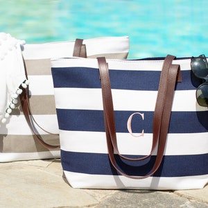 May include: Two striped canvas tote bags with brown leather handles. One bag is gray and white, the other is navy blue and white. The navy blue bag has a pink letter "C" embroidered on the front.