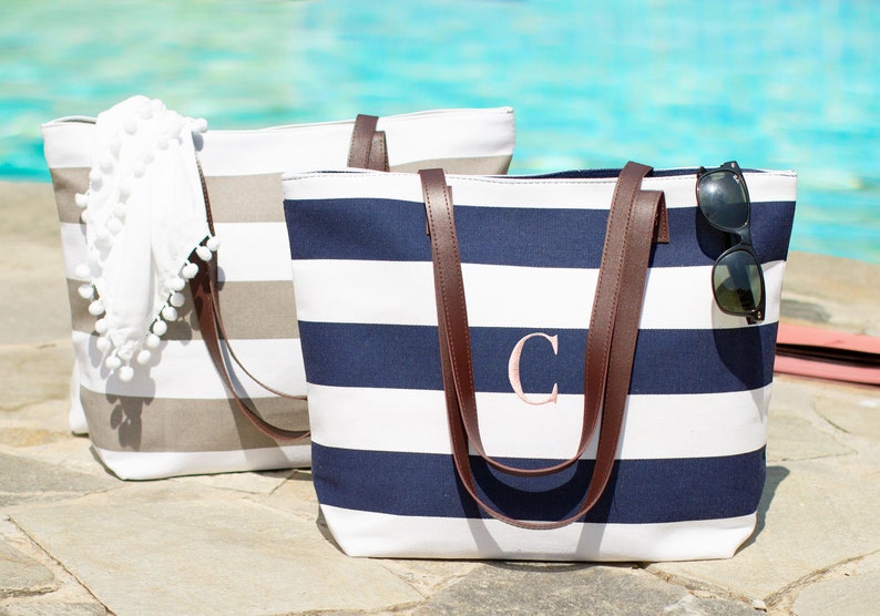 May include: Two striped canvas tote bags with brown leather handles. One bag is gray and white, the other is navy blue and white. The navy blue bag has a pink letter "C" embroidered on the front.