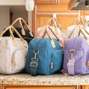 May include: A collection of corduroy travel bags in various colors, including black, pink, teal, lavender, and cream. Each bag features a top handle, a shoulder strap, and gold-colored hardware. The bags are displayed on a countertop.