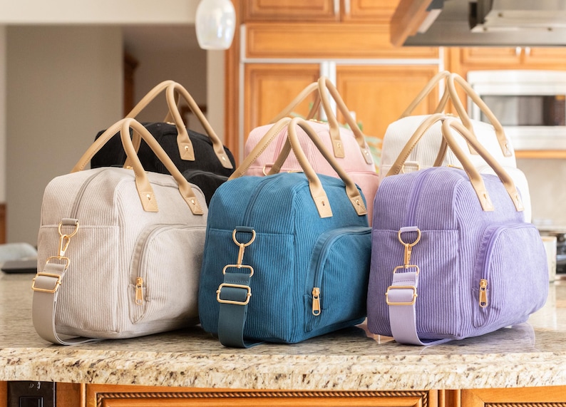 May include: A collection of corduroy travel bags in various colors, including black, pink, teal, lavender, and cream. Each bag features a top handle, a shoulder strap, and gold-colored hardware. The bags are displayed on a countertop.