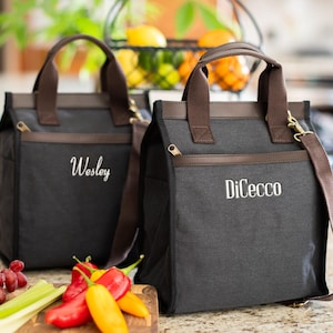 May include: Two black insulated lunch bags with brown leather trim and handles. One bag has the name "Wesley" embroidered in white, the other has "DiCecco". Both bags have a zippered top, side pockets, and a detachable shoulder strap. A cutting board with vegetables is in the foreground.