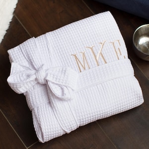 May include: A white waffle-weave robe featuring a tied waist belt and gold-colored embroidered monogram letters MKE on the chest area. The fabric has a distinct grid-like texture, and the garment is folded neatly into a compact rectangular shape.