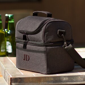May include: A gray insulated lunch bag with a black zipper and a black shoulder strap. The bag has a personalized monogram "JD" on the front.