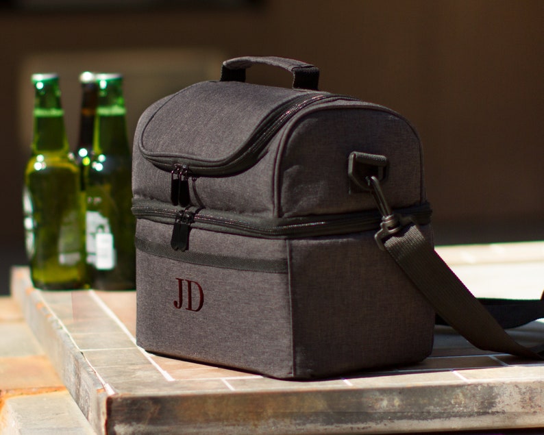 May include: A gray insulated lunch bag with a black zipper and a black shoulder strap. The bag has a personalized monogram "JD" on the front.