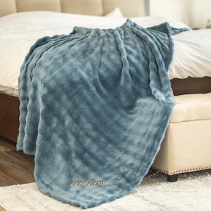 May include: A soft, blue blanket draped over a bed. The blanket has a textured surface and is embroidered with the Roman numerals VII.XXVI.XXI. The bed has a white comforter and is next to a beige bench.