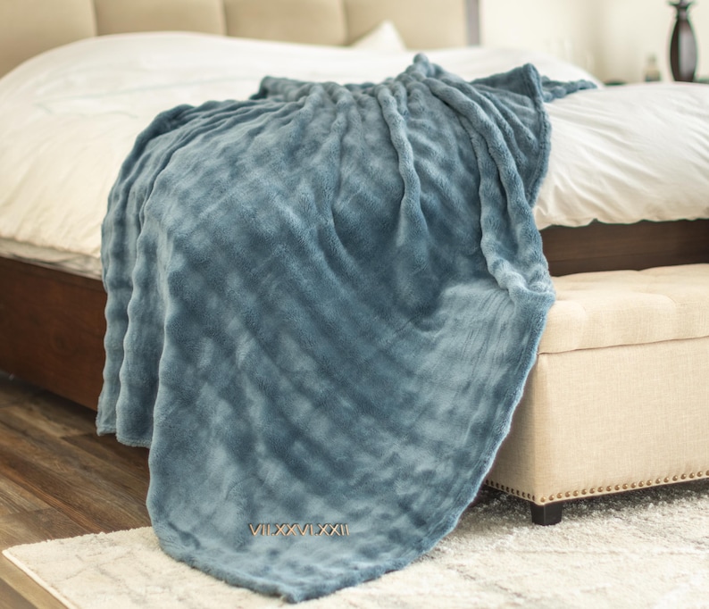 May include: A soft, blue blanket draped over a bed. The blanket has a textured surface and is embroidered with the Roman numerals VII.XXVI.XXI. The bed has a white comforter and is next to a beige bench.