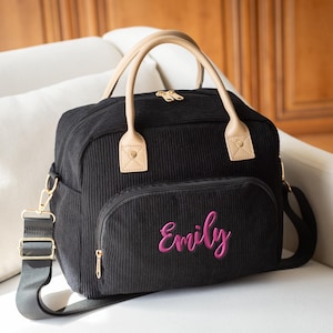 May include: Black corduroy tote bag with a front pocket and a shoulder strap. The bag has tan handles and gold-colored hardware. The name "Emily" is embroidered in pink on the front pocket.