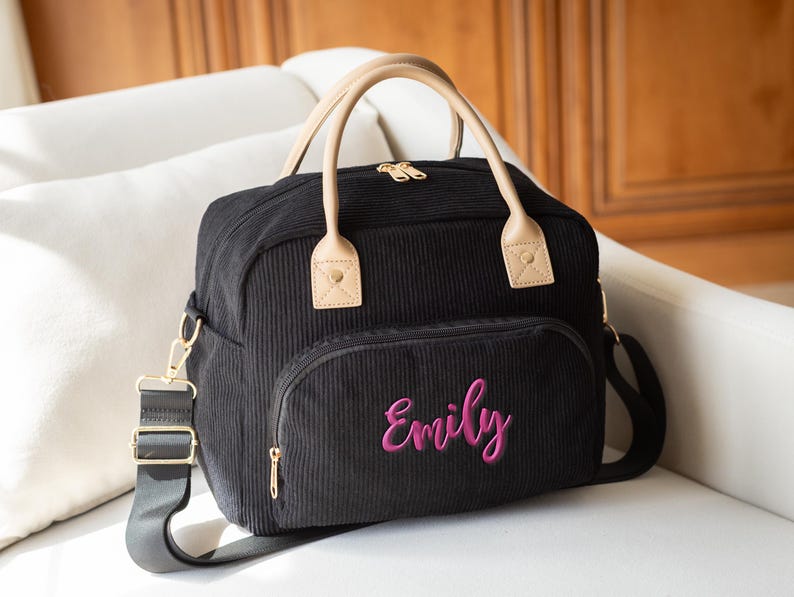 May include: Black corduroy tote bag with a front pocket and a shoulder strap. The bag has tan handles and gold-colored hardware. The name "Emily" is embroidered in pink on the front pocket.