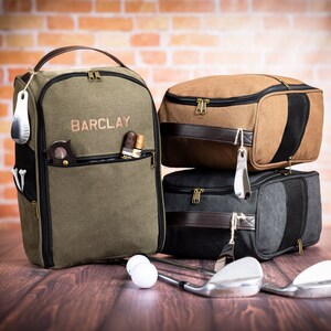 May include: A green canvas golf bag with the name "BARCLAY" embroidered on the front. The bag is sitting on a wooden surface with two brown and black golf bags stacked on top of each other. There are golf balls and golf clubs in the image.