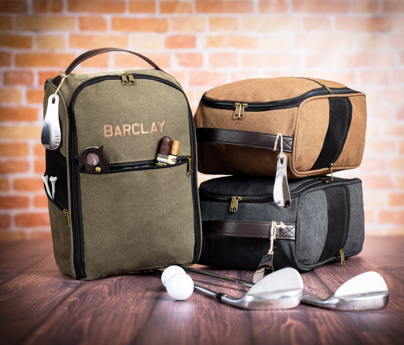 May include: A green canvas golf bag with the name "BARCLAY" embroidered on the front. The bag is sitting on a wooden surface with two brown and black golf bags stacked on top of each other. There are golf balls and golf clubs in the image.
