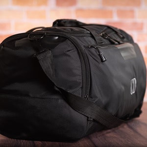 K&ouml;nnte beinhalten: Schwarze Reisetasche mit Rei&szlig;verschlussfach und schwarzem Trageriemen. Die Tasche hat ein kleines Logo an der Seite.