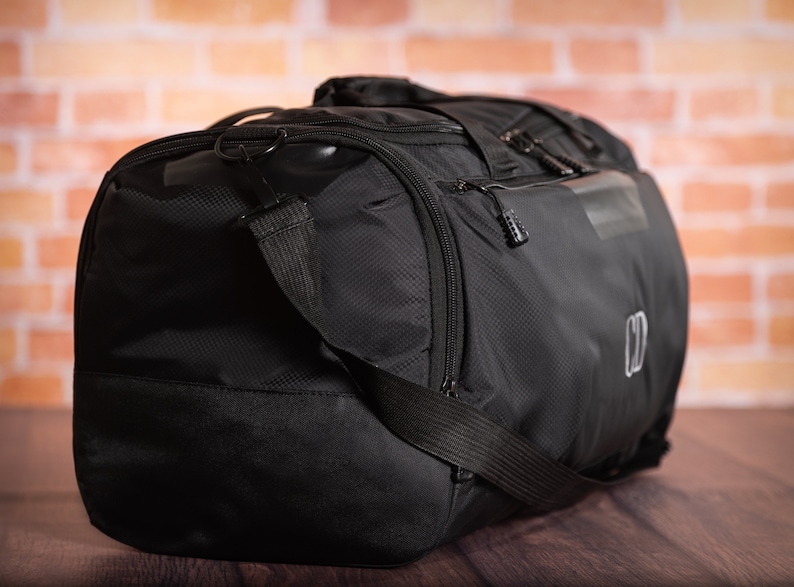 K&ouml;nnte beinhalten: Schwarze Reisetasche mit Rei&szlig;verschlussfach und schwarzem Trageriemen. Die Tasche hat ein kleines Logo an der Seite.