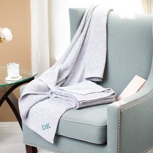 May include: A gray throw blanket with the initials "DK" embroidered on it is draped over a light blue armchair. The chair has a tufted back and nailhead trim.