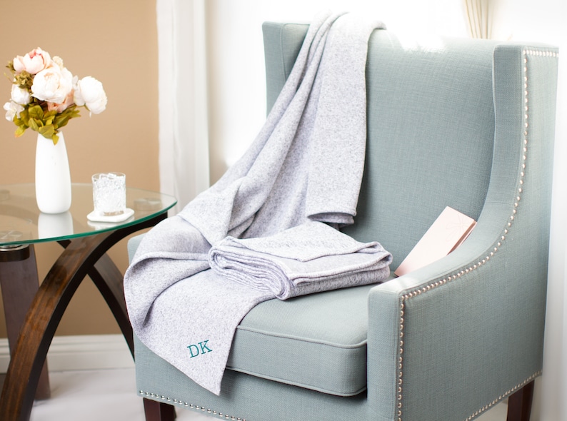 May include: A gray throw blanket with the initials "DK" embroidered on it is draped over a light blue armchair. The chair has a tufted back and nailhead trim.