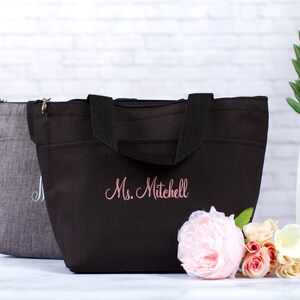 May include: A black insulated lunch bag with a zipper closure and two black handles. The bag has a pink embroidered name "Ms. Mitchell" on the front.