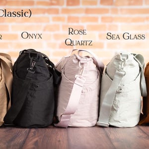 May include: Five classic style shoulder bags in various colors: Sandbar, Onyx, Rose Quartz, Sea Glass, and Ember. Each bag has a matching adjustable shoulder strap and a side pocket. The bags are displayed against a brick wall.