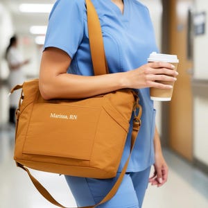 May include: A mustard-colored shoulder bag with the name "Marissa, RN" printed on the front. The bag has a long strap and is carried by a person wearing blue scrubs. A disposable coffee cup is held in the person's hand.