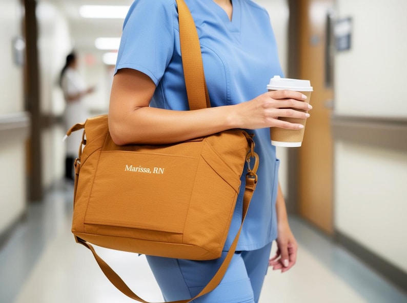 May include: A mustard-colored shoulder bag with the name "Marissa, RN" printed on the front. The bag has a long strap and is carried by a person wearing blue scrubs. A disposable coffee cup is held in the person's hand.