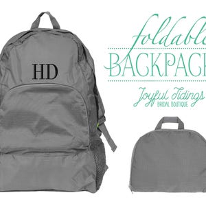 May include: A gray foldable backpack with the initials "HD" embroidered on the front. The backpack is folded and laid flat next to a smaller gray foldable backpack.
