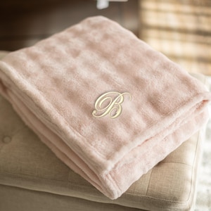 May include: A folded, light pink plush blanket with a gold-colored embroidered letter "B" in a script font. The blanket is resting on a beige upholstered ottoman. The soft blanket is a cozy home decor item.