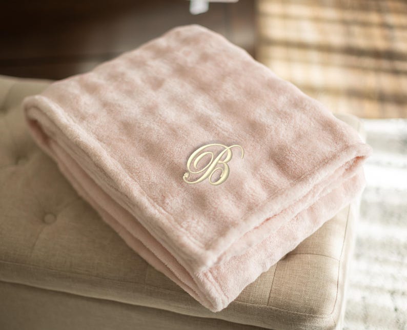 May include: A folded, light pink plush blanket with a gold-colored embroidered letter "B" in a script font. The blanket is resting on a beige upholstered ottoman. The soft blanket is a cozy home decor item.
