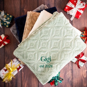 May include: A stack of soft, plush blankets in various colors, including green, beige, and white. The top blanket is green with a diamond pattern and the text "Gigi est 2024" embroidered in green.