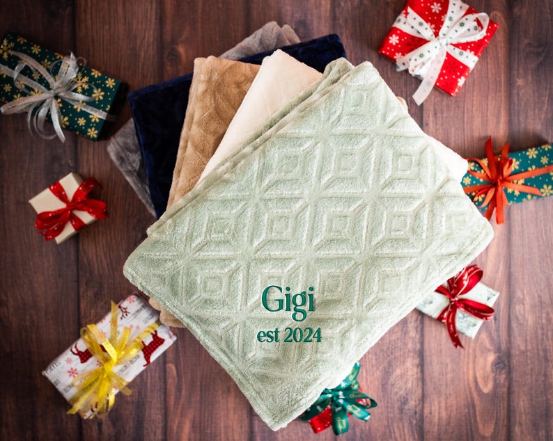 May include: A stack of soft, plush blankets in various colors, including green, beige, and white. The top blanket is green with a diamond pattern and the text "Gigi est 2024" embroidered in green.
