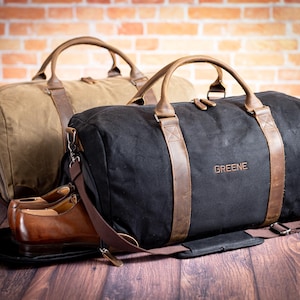 May include: Two canvas duffel bags with leather accents and straps. One bag is tan and the other is black. The black bag has the name "Greene" embroidered on it.